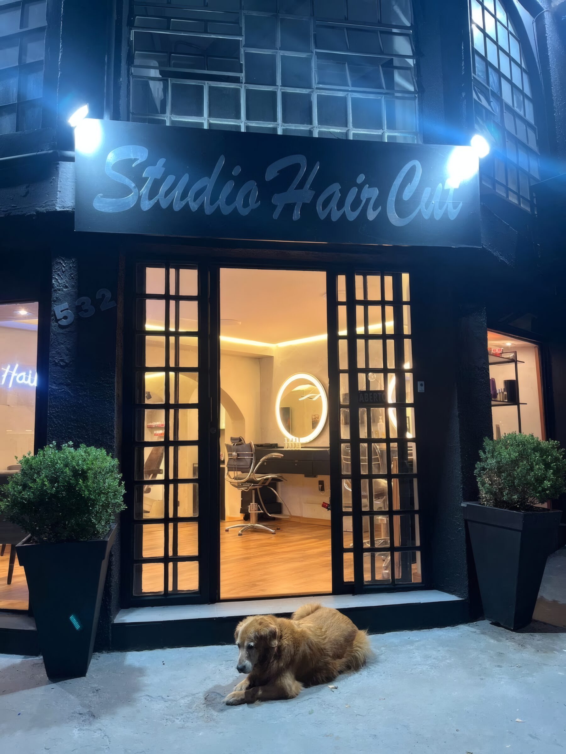 Fachada preta do Studio Hair Cut à noite, com a luz acesa por dentro e um cachorro deitado na porta